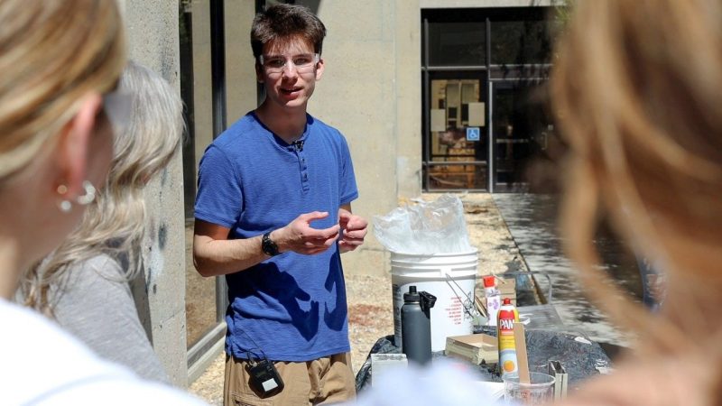 A student in safety goggles, standing at a table outside, speaks to a group of a people. 