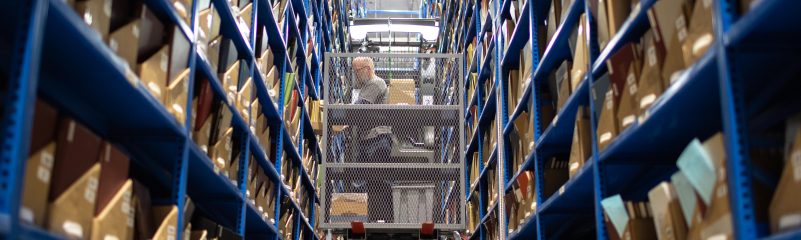A man standing in a mobile elevated work platform, or boom lift, grabs a box of books from tall blue shelves.